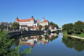 Schloss Neuburg und die Donau, die sich rechts am Schloss vorbeischlängelt. Links ist die Promenade und links das begrünte Ufer zu sehen.