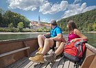 In dem hölzernen Boot sitzen ein Mann und eine Frau, beide in Wanderkleidung mit dem Blick Richtung Kloster Weltenburg gerichtet. Das Kloster ist im Hintergrund von weitem zu sehen umrahmt von grünen Wäldern.