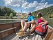 In dem hölzernen Boot sitzen ein Mann und eine Frau, beide in Wanderkleidung mit dem Blick Richtung Kloster Weltenburg gerichtet. Das Kloster ist im Hintergrund von weitem zu sehen umrahmt von grünen Wäldern.