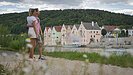 Ein Pärchen in sommerlicher Kleidung steht am Main-Donau-Kanal und halten sich im Arm. Sie schauen aufs Flussufer gegenüber, dort ist die Riedenburger Promenade zu sehen. Dahinter ragen die Hänge mit ihrem dunkelgrünen Kleid auf.