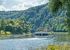 Ein Personenschiff fährt durch die Weltenburger Enge am Donaudurchbruch. Die Wälder an den Ufern erstrahlen in leuchtend grünen Farben. Der Himmel blitzt blau durch.