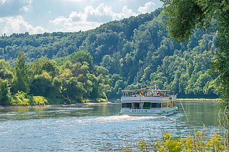 Schifffahrt durch die Weltenburger Enge Ein Personenschiff fährt durch die Weltenburger Enge am Donaudurchbruch. Die Wälder an den Ufern erstrahlen in leuchtend grünen Farben. Der Himmel blitzt blau durch.