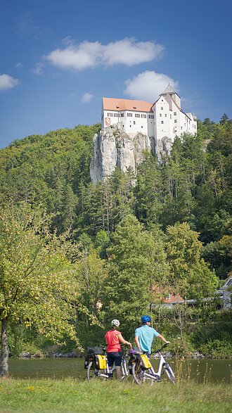Radler unterwegs auf dem Altmühltal-Radweg bei Burg Prunn Zwei Radler mit Fahrradhelmen, sowie rotem und blauen Shirt stehen am Radweg. Ihre Fahrräder halten sie auf der rechten Seite, beide haben gelb-schwarze Satteltaschen. Die zwei Radler schauen über den Main-Donau-Kanal hinauf zur Burg Prunn, die umgeben von Wäldern auf einem Felsen hochoben sitzt. Der Himmel scheint blau, nur ein paar Wolken verirren sich.
