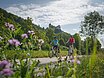 Zwei Radler, eine Frau mit weißem Helm und roten Shirt und ein Mann, neben ihr, ebenfalls mit Helm und blauen Shirt sind auf dem Radweg unterwegs. Die Zwei radeln und hinter ihnen ragt die Burg Prunn, die umgeben von Wäldern auf einem Felsen. Der Himmel scheint blau, nur ein paar Wolken verirren sich.