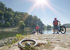 Zwei Radler mit Fahrrädern stehen am Ufer der Donau und warten auf die Fähre, die sie ans andere Ufer bringen wird.