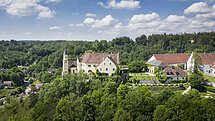 Das Schloss Hexenagger von oben zu sehen. Eingebettet in grünem Blätterkleid. Der Himmel ist hellblau und weiße Wolken sind zu sehen.